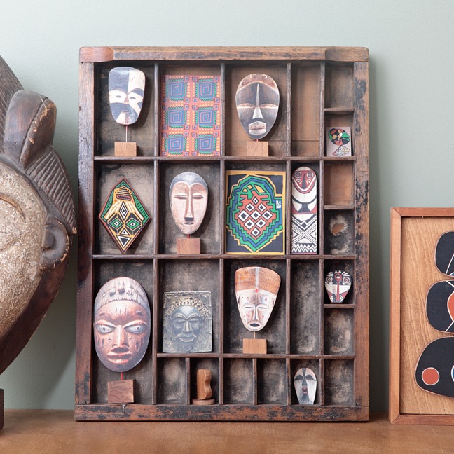 Handmade African mask ornaments and other African themed original artworks and photography displayed in an old antique letterpress printers tray type case drawer.